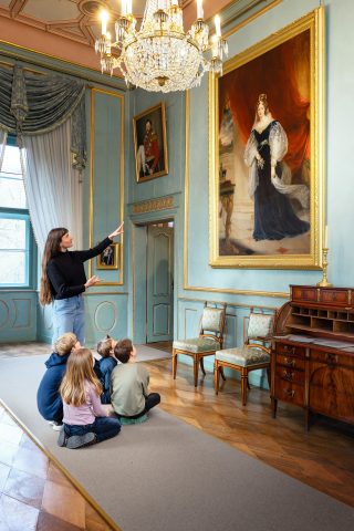Eine junge Frau zeigt auf ein Gemälde von Prinzessin Adelheid von Sachsen‑Meiningen, während Kinder auf dem Boden sitzen und aufmerksam zuhören.