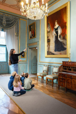 Eine junge Frau zeigt auf ein Gemälde von Prinzessin Adelheid von Sachsen‑Meiningen, während Kinder auf dem Boden sitzen und aufmerksam zuhören.