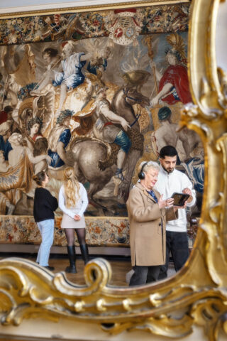 Zwei Frauen betrachten die Alexandertapisserie, während ein junger Mann und eine Frau auf einem digitalen Guide Informationen ansehen; die Frau trägt Kopfhörer.