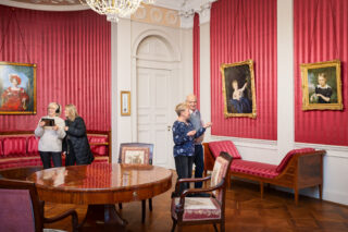 Besucherinnen und Besucher in einem historischen Raum des Schlosses Elisabethenburg; einige betrachten Kunstwerke, andere nutzen einen digitalen Guide.
