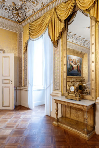 Prunkvoller Raum mit goldverziertem Spiegel, antiker Uhr auf einem goldenen Konsolentisch und großen Fenstern mit goldenen Vorhängen.