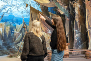 Zwei Frauen stehen vor einem historischen Bühnenbild im Theatermuseum; eine der Frauen zeigt mit dem Finger darauf.